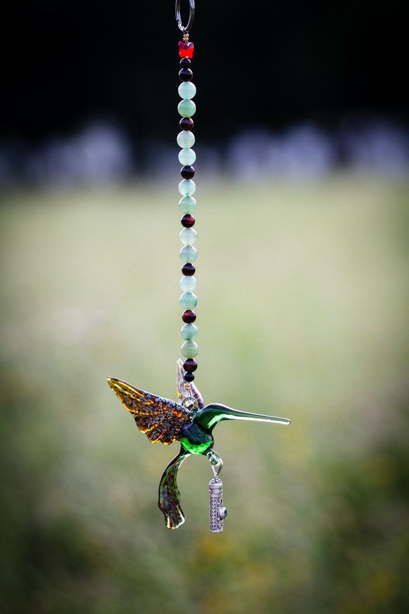 Green and tan glass hummingbird charm with a crystal butterfly, suspended by a beaded strand and a silver keepsake urn pendant.