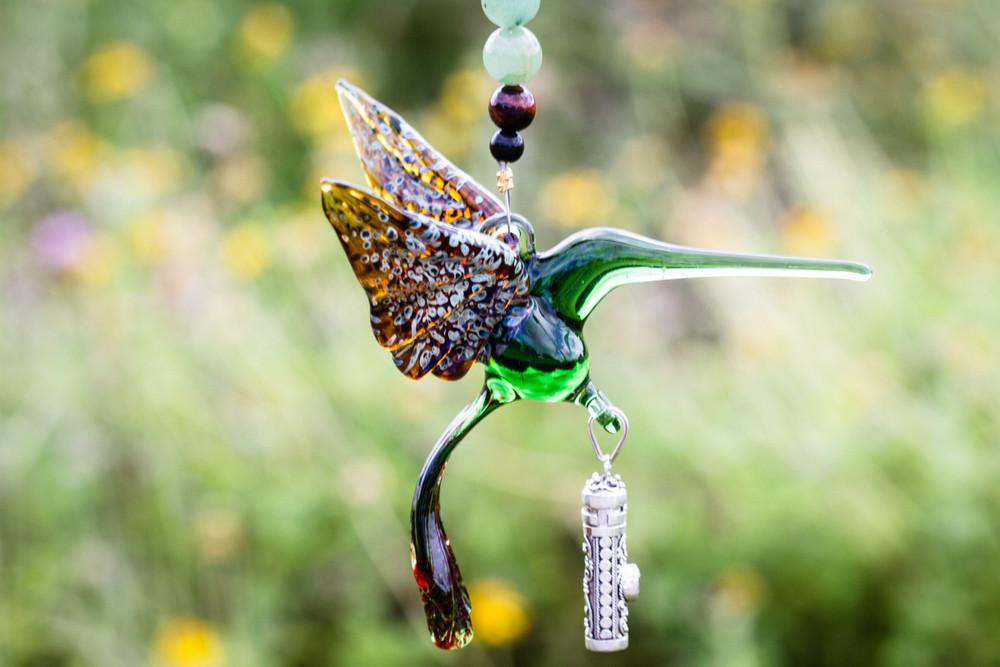 Green glass hummingbird with outstretched wings and a dangling ornate silver keepsake urn pendant against a sunny field.