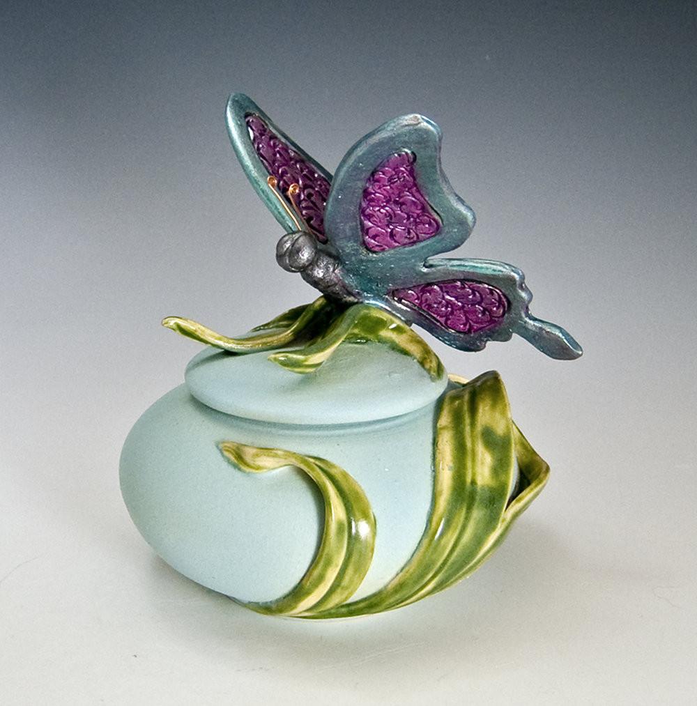 Handmade butterfly ceramic urn with purple winged butterfly perched on a pale blue jar wrapped in green leaves.