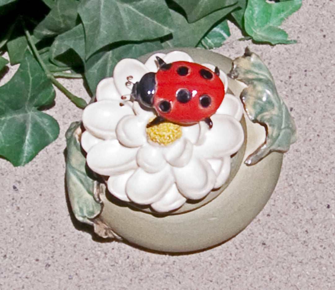 Handmade ladybug ceramic keepsake urn resting on a white flower with a yellow center and green leaves