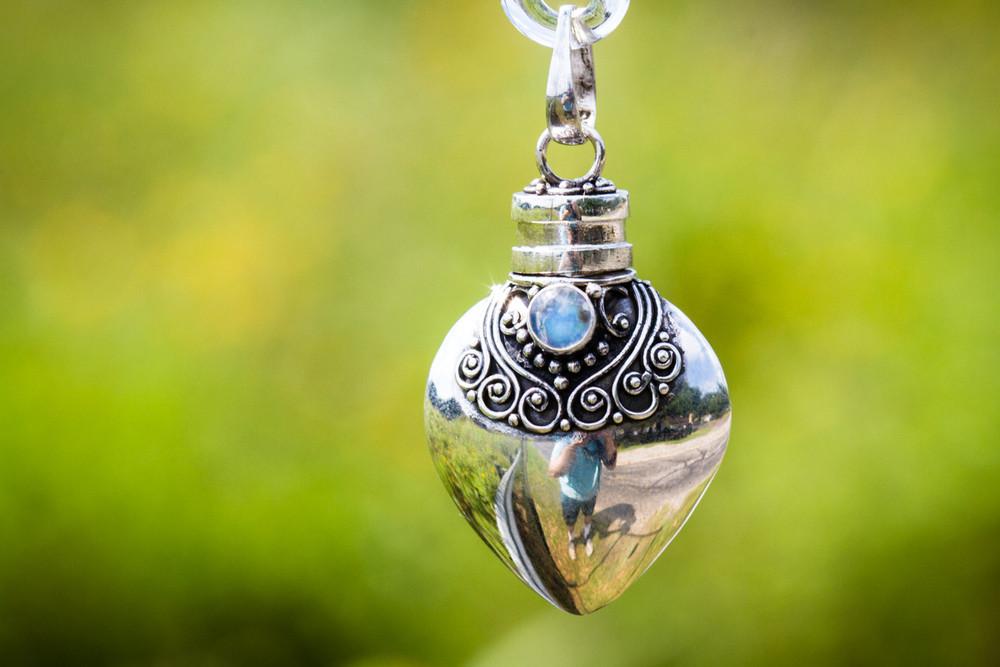 Close-up of a polished sterling silver heart keepsake urn pendant with intricate filigree and a blue gemstone against a green background.