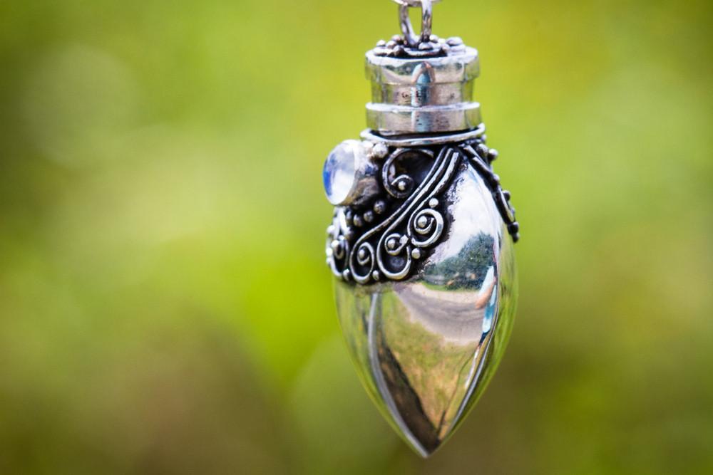 Close-up of the ornate sterling silver heart keepsake urn pendant with filigree and a blue stone, reflecting a bright outdoor scene.