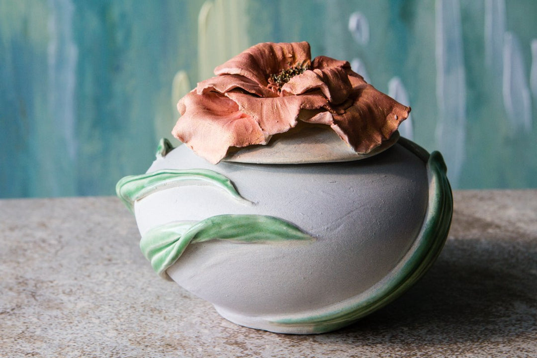 Handsculpted ceramic urn for cremated ashes with a large pink bloom and green leaf accents.