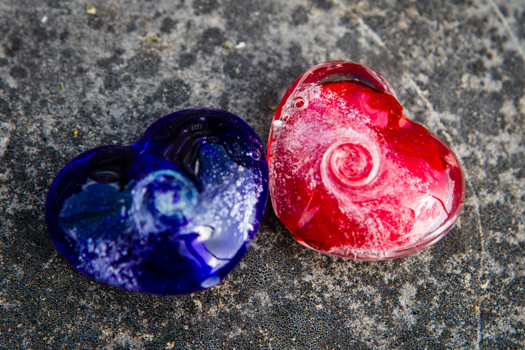 Two heart shaped glass cremains charms in blue and red on a textured rock surface.