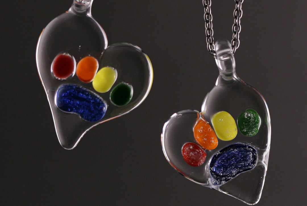 Two glass Rainbow Bridge pawprint pendants filled with bright colored beads on a dark background