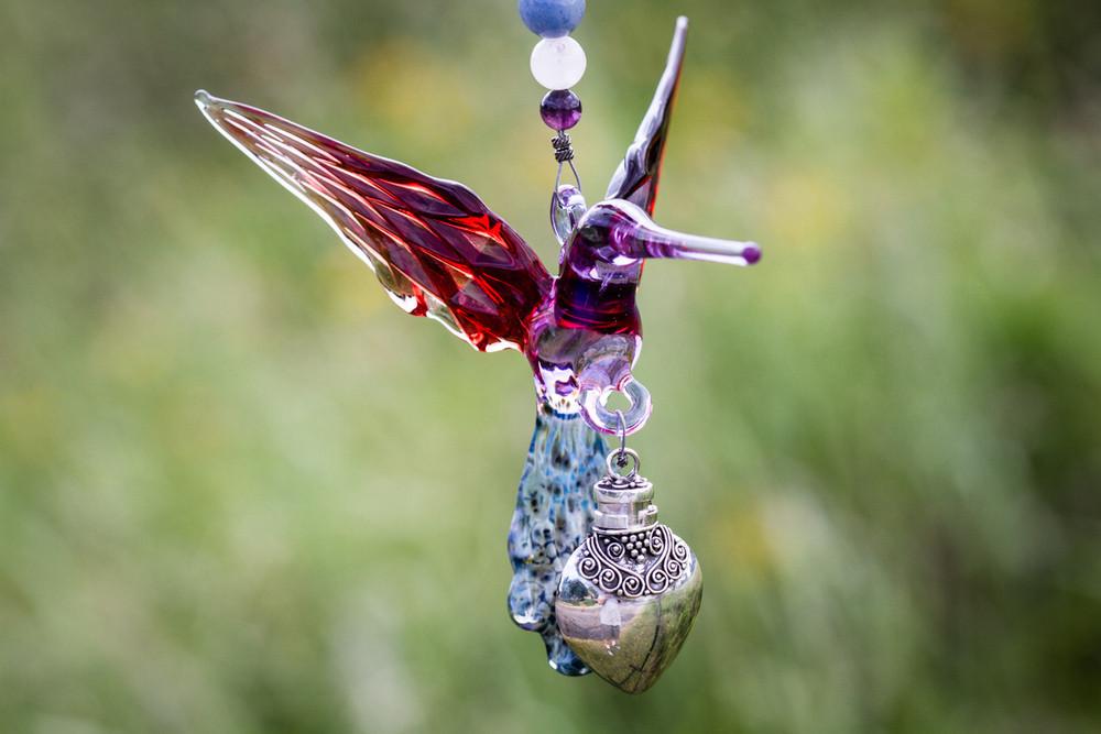 Handmade glass hummingbird with purple body and red wings, suspended by a beaded string with a silver keepsake urn pendant.