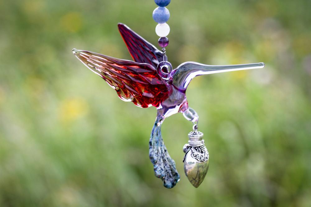 Close-up of a handmade glass hummingbird with purple body and red wings, hanging from a beaded string with a silver keepsake urn pendant.