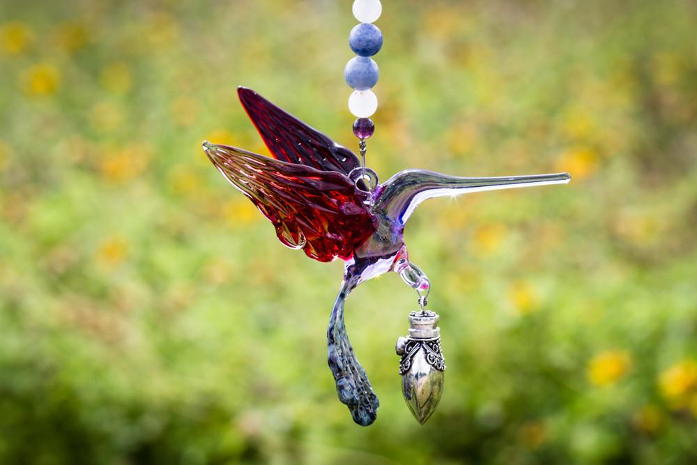 Red and purple glass hummingbird ornament with a silver keepsake urn pendant and beaded strand against a blurred garden background