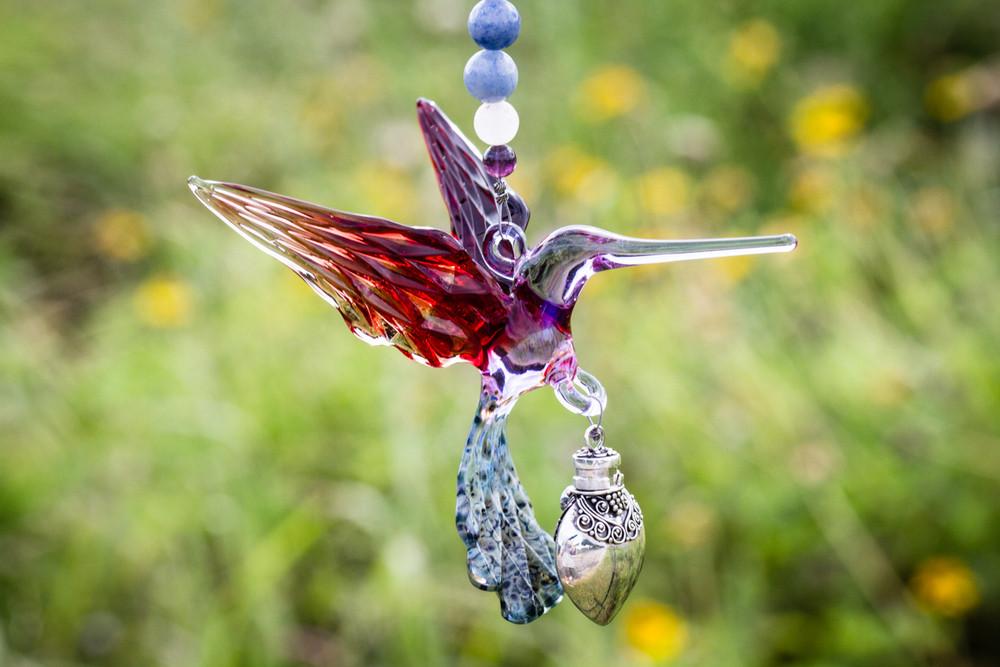 Purple and red glass hummingbird charm with a silver keepsake urn and beaded accents hanging in front of pink flowers