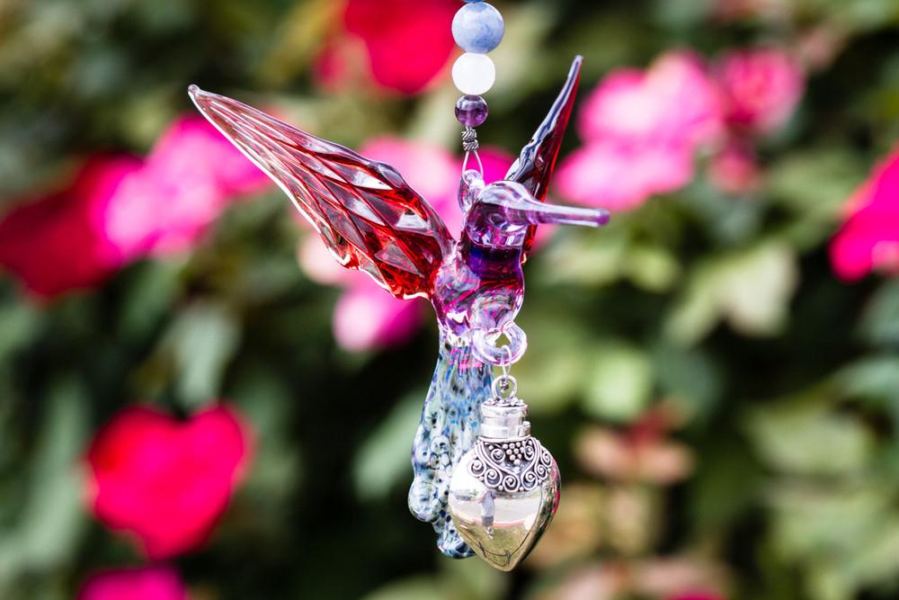 Side view of red and purple glass hummingbird with an ornate silver keepsake urn and bead chain outdoors among blooming plants