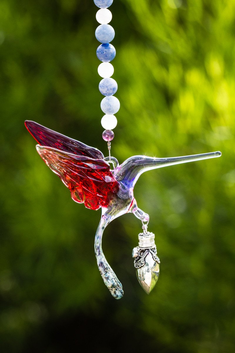 Glass hummingbird pendant with red and light purple wings hanging from a blue and white bead strand with a keepsake vial.