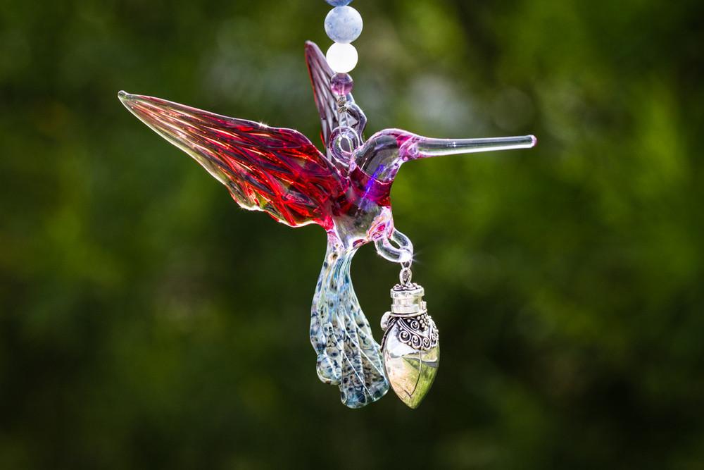 Glass hummingbird pendant with red and purple wings and an ornate silver keepsake vial charm on a bead necklace.