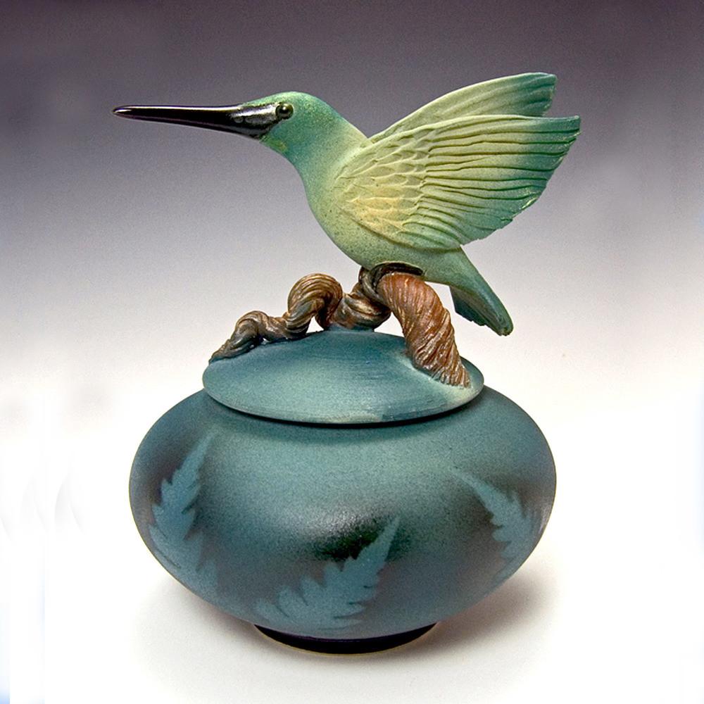 Handmade ceramic urn jar for ashes featuring a green hummingbird perched on a branch atop a blue round vessel.