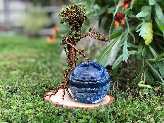 Ent wire sculpture outdoors with blue orb on wooden base in a garden setting