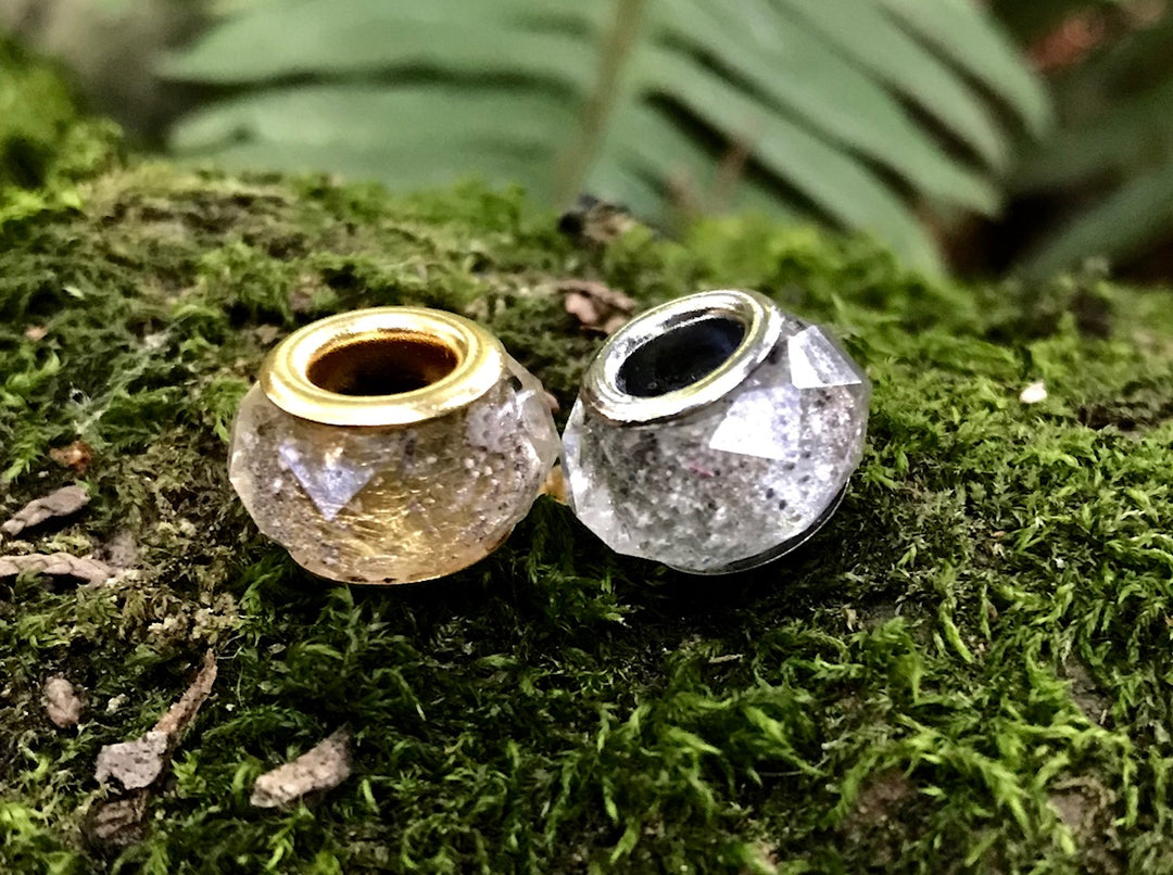 Two crystal-cut cremains beads with gold and silver inner rims rest on mossy rocks.