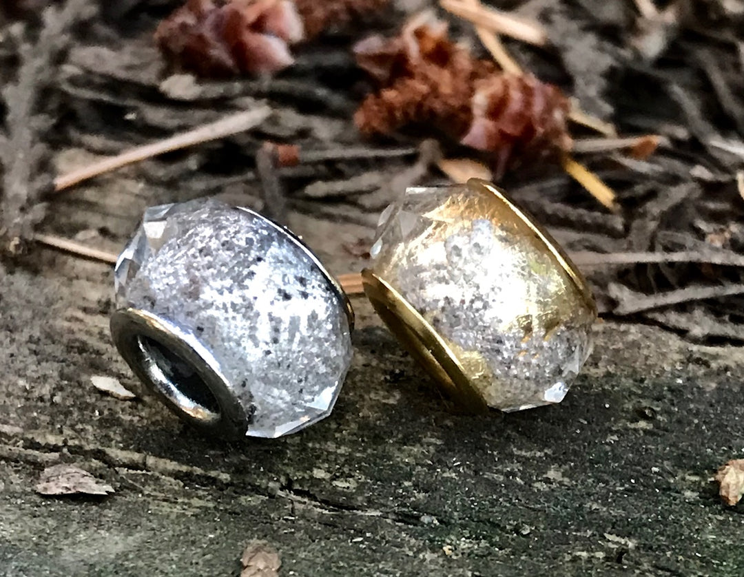 Two crystal-cut cremains beads in silver and gold tones resting on a rough outdoor surface.