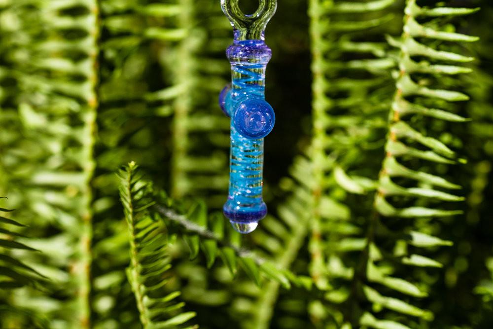 Blue and purple glass cross pendant infused with cremains, shown from the side with decorative cap against greenery.
