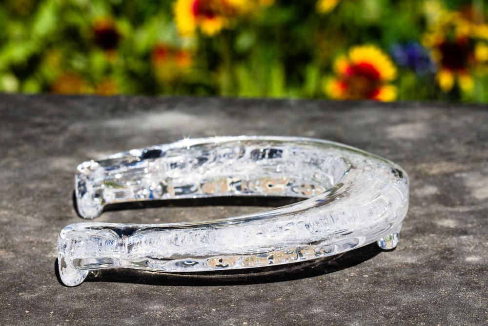 Clear glass horseshoe cremation sculpture resting on a stone surface with a colorful garden background.