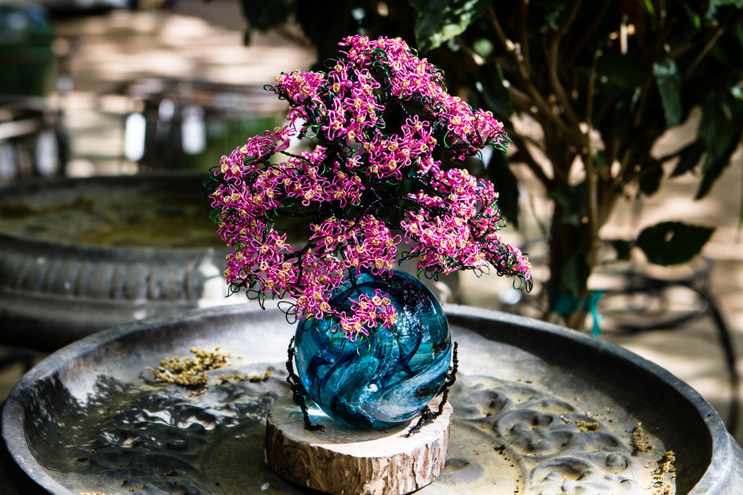Pink dogwood tree on teal rope orb perched on a natural wood base in a garden.