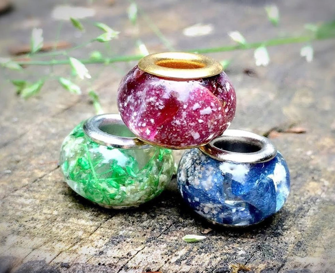 Three speckled cremains beads in green pink and blue with silver cores laid on a rough surface.