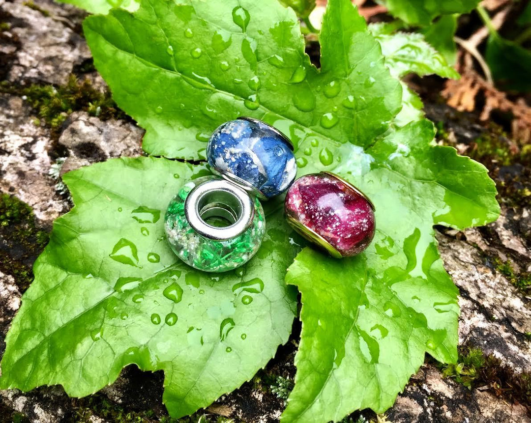 Three Pandora compatible cremains beads for keepsakes in blue green and purple on a mossy leaf