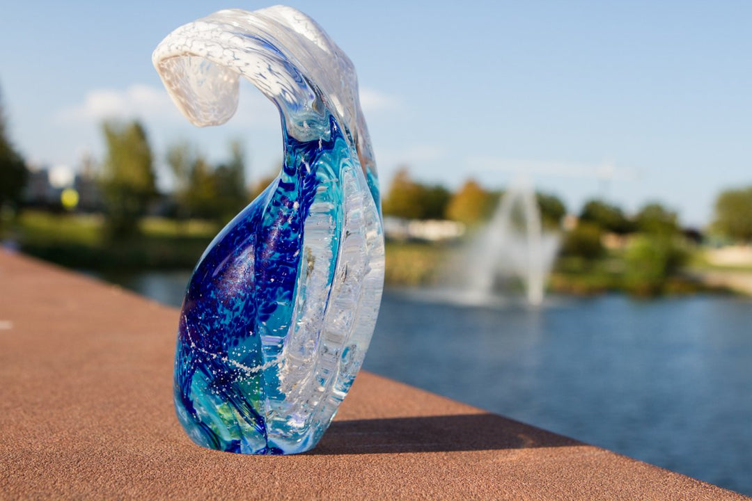 Blue and clear glass ocean wave paperweight on a brick ledge with a park and fountain in the background
