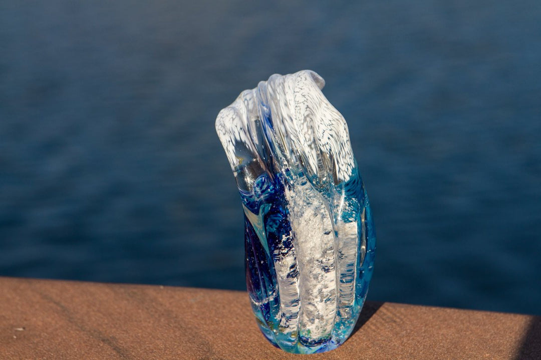 Sculpted glass ocean wave paperweight with blue and clear swirls resting on a ledge beside the deep blue ocean