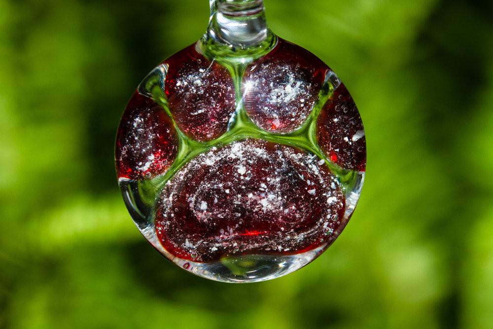 Paw print glass pendant with red hues and white speckles infused with cremains against a green backdrop