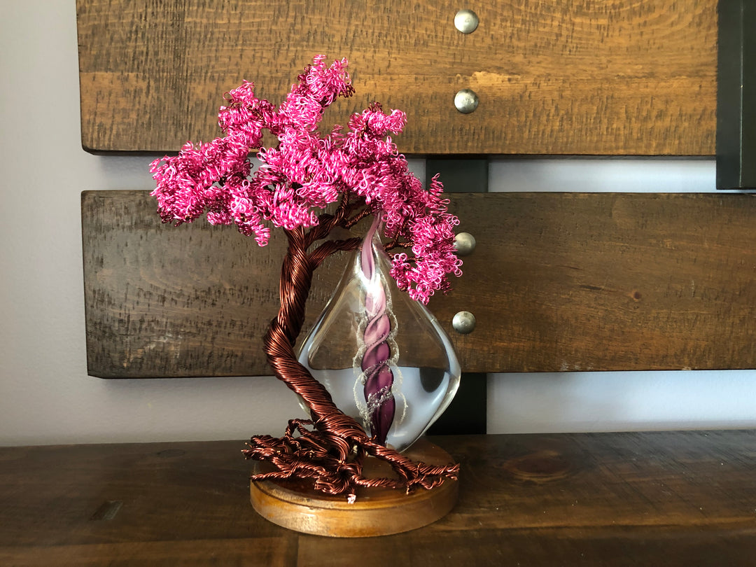 Pink wire Tree of Life with curly blossoms and purple spiral flame inside a glass teardrop vase on a wooden base.