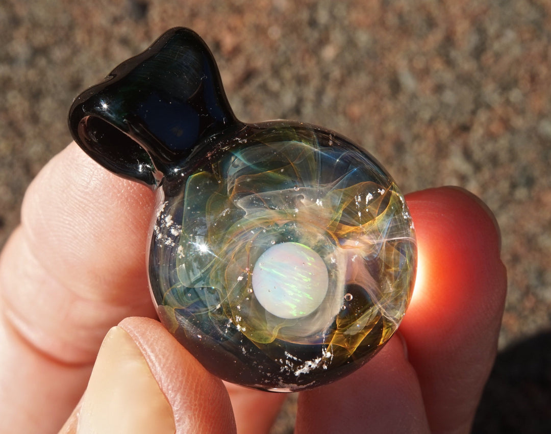 Handheld glass planet pendant with opal core and cremains, swirling gold and silver fumes, dark bail at top.