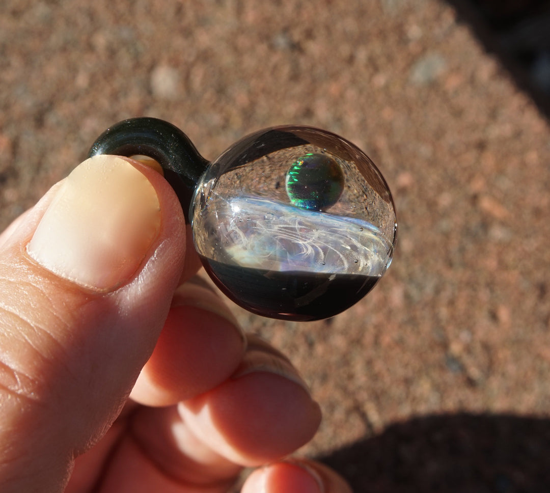Close-up of a glass planet pendant with a green opal inside a clear swirled core and a dark curved bail held against a rocky ground.