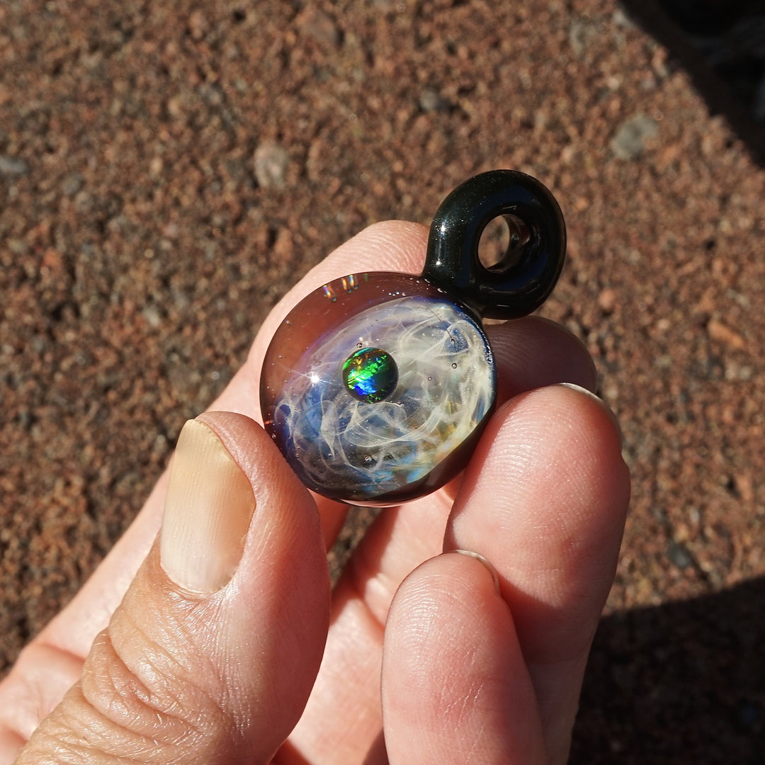 Close-up of a glass planet pendant with an iridescent opal and wispy interior, held between fingers outdoors.