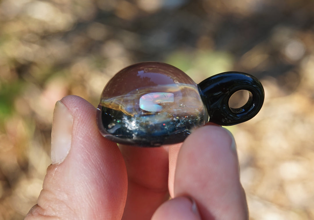Close-up of a glass planet pendant with a vivid opal and swirling interior set in a dark brown outer shell held by fingers.