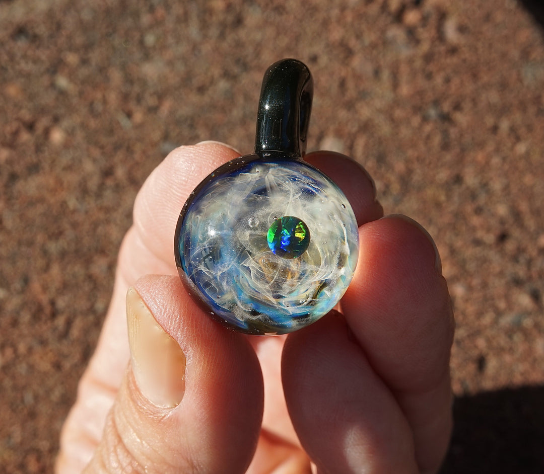 Round galaxy glass pendant with blue white swirls and a green opal center, held between fingers by a dark bail