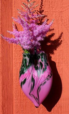 Purple swirl hanging vase with cremains, displayed on an orange wall with pink flowering stems.