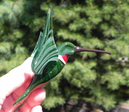 Side view of cremation ashes infused green glass hummingbird with red throat against a leafy background.