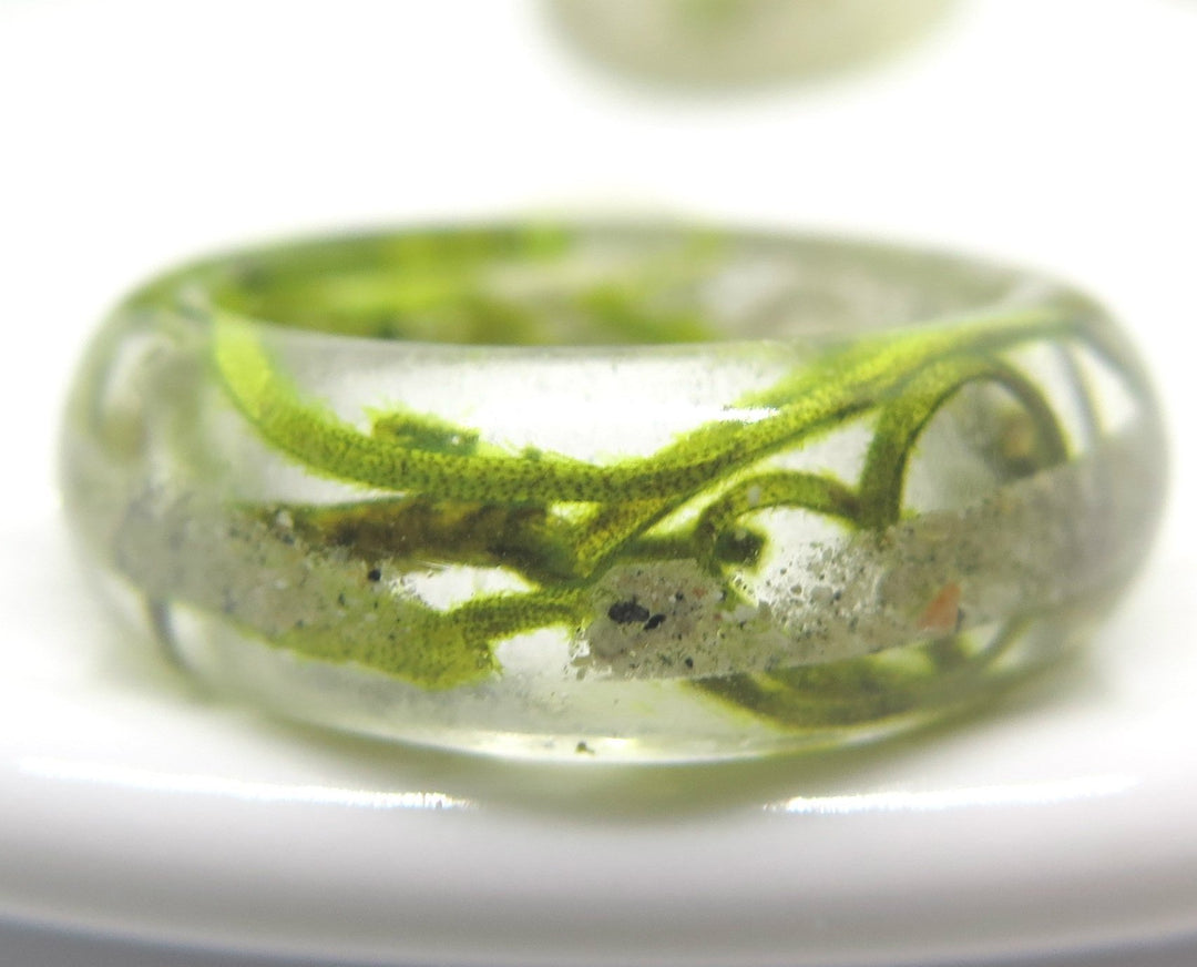 Green moss ring in clear resin with embedded ash and moss strands resting on a white surface.