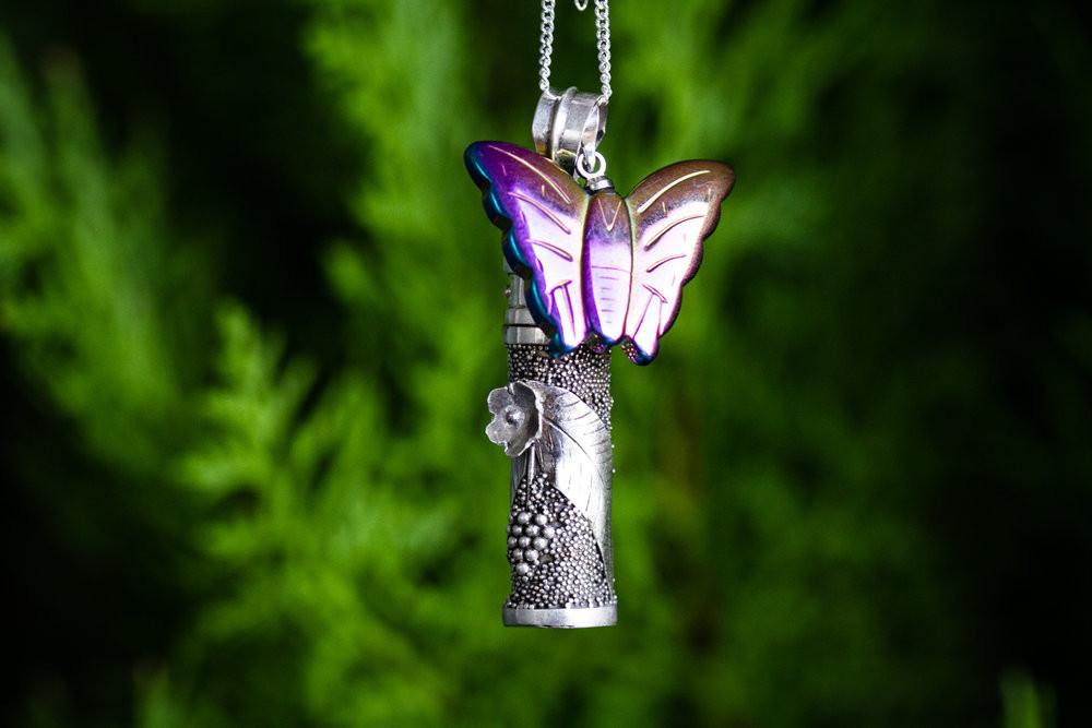 Silver cremation necklace with a textured cylindrical pendant and a purple butterfly perched on top against green foliage
