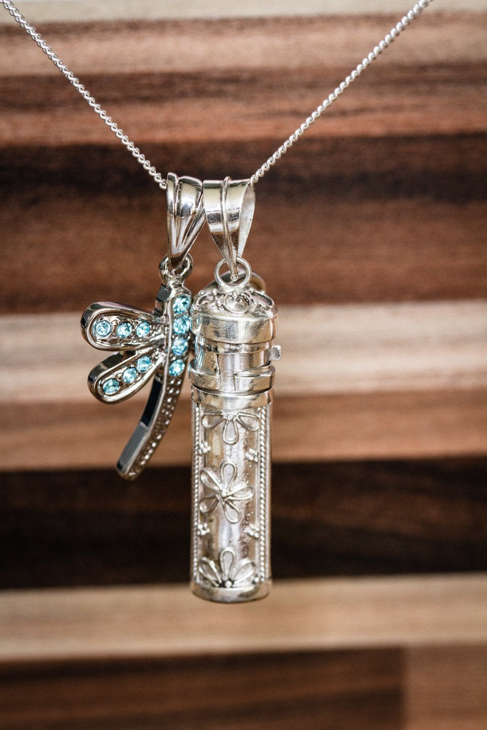 Close-up of sterling silver keepsake necklace with a turquoise crystal dragonfly charm and the floral engraved cremation capsule pendant.