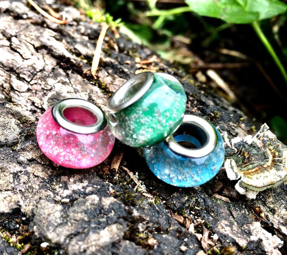 Pink cremation bead with a silver metal core resting on a wooden surface.