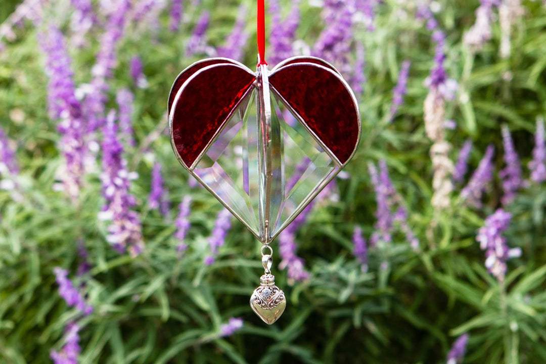 Red stained glass memorial heart suncatcher with a silver keepsake pendant against a backdrop of purple flowers.