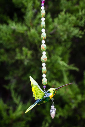 Hummingbird ornament suspended by a beaded necklace with a decorative heart keepsake pendant against a garden backdrop.