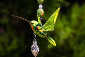 Green glass hummingbird charm with a sterling silver heart keepsake urn and beaded necklace against a blurred green background.