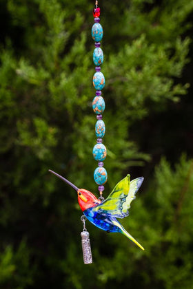 Vertical strand of turquoise marbled beads with purple spacers supporting a colorful glass hummingbird and a small silver keepsake pendant