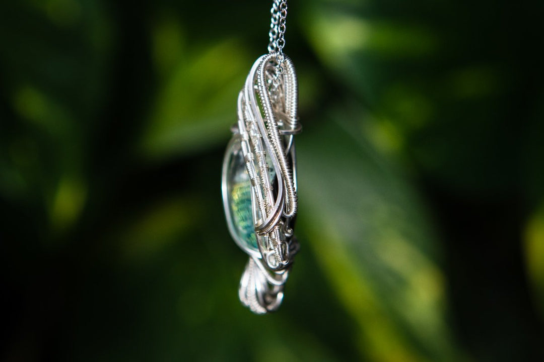 Close up of a silver wire wrapped Madison pendant with a dichroic cabochon containing cremation ashes, suspended on a chain.