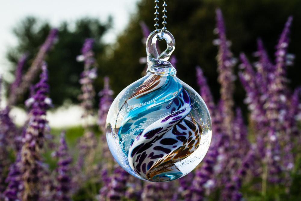 Close up of a multicolored glass cremation orb with blue purple brown white swirls hanging from a metal chain against a garden background.