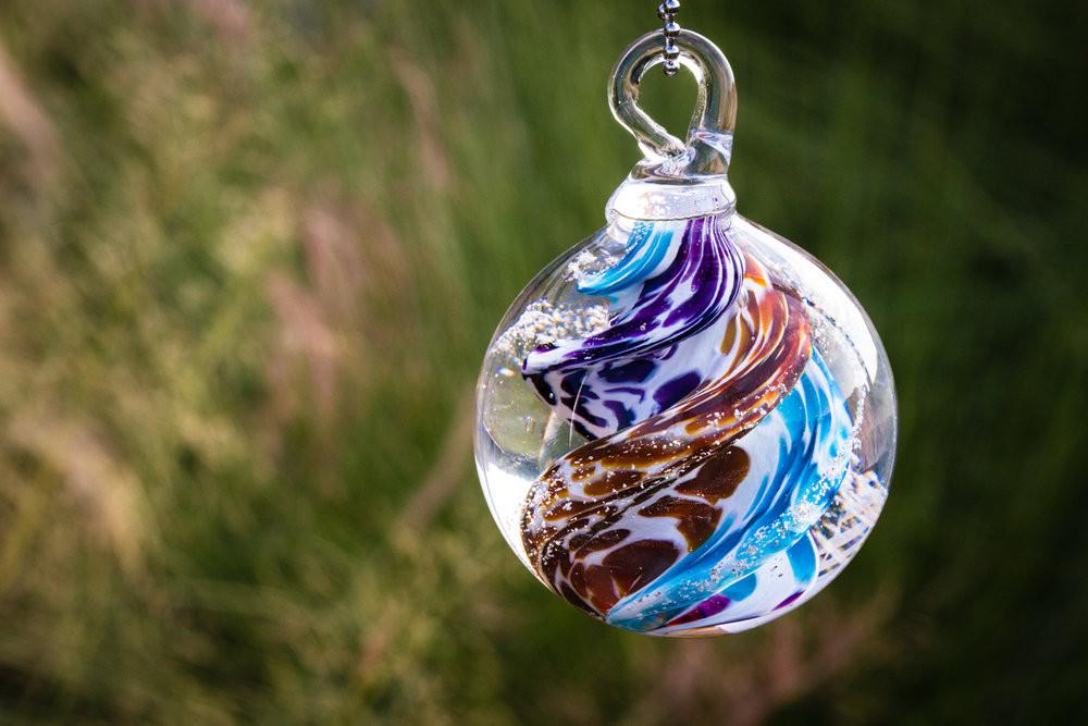 Close view of a swirled glass cremation orb in blue brown purple and white suspended by a loop against a green outdoor backdrop.