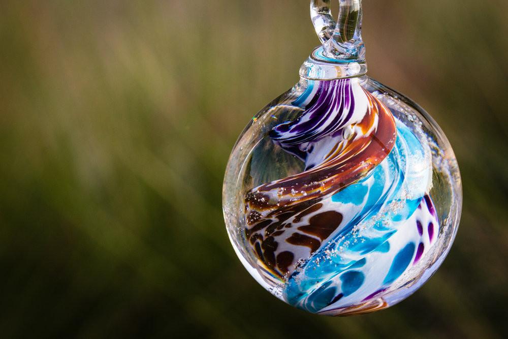 Tan and purple swirled cat eye glass cremation orb with cremains, hanging from a loop in a garden or home setting.