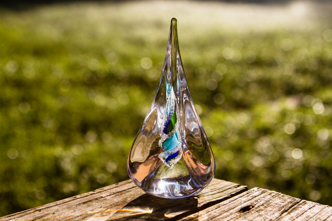 Tri-color glass memorial flame with cremation ash visible in a spiral, displayed outdoors on a wooden surface.