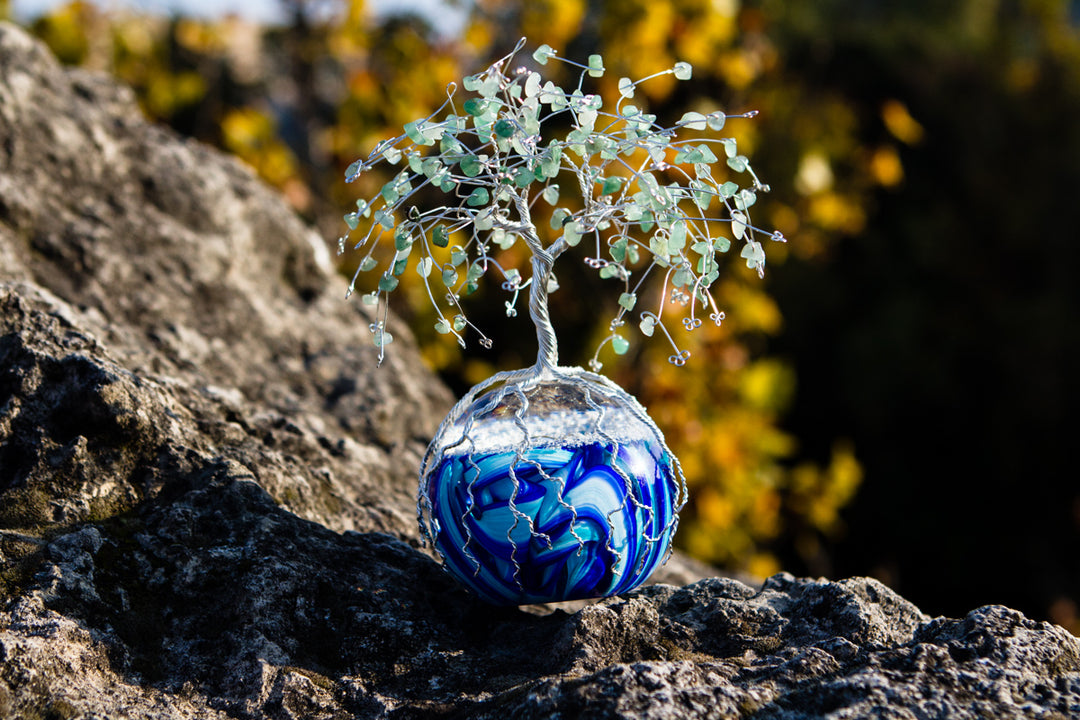 Blue swirled glass orb with a silver wire tree of life and green aventurine beads on a rocky surface.